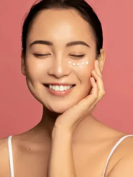 A smiling woman with even skin tone applies some skin care cream beneath her eyes for glowing skin.