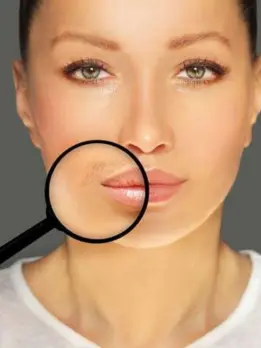 A woman using a magnifying lens is looking at her face & closely checking the skin on her upper lip.