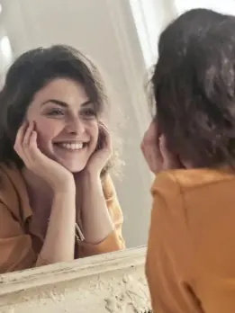 After melasma skin care treatment, a happy woman in an orange outfit looks at herself in the mirror