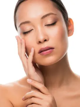 A smiling woman with even skin tone applies some skin care cream beneath her eyes for glowing skin.